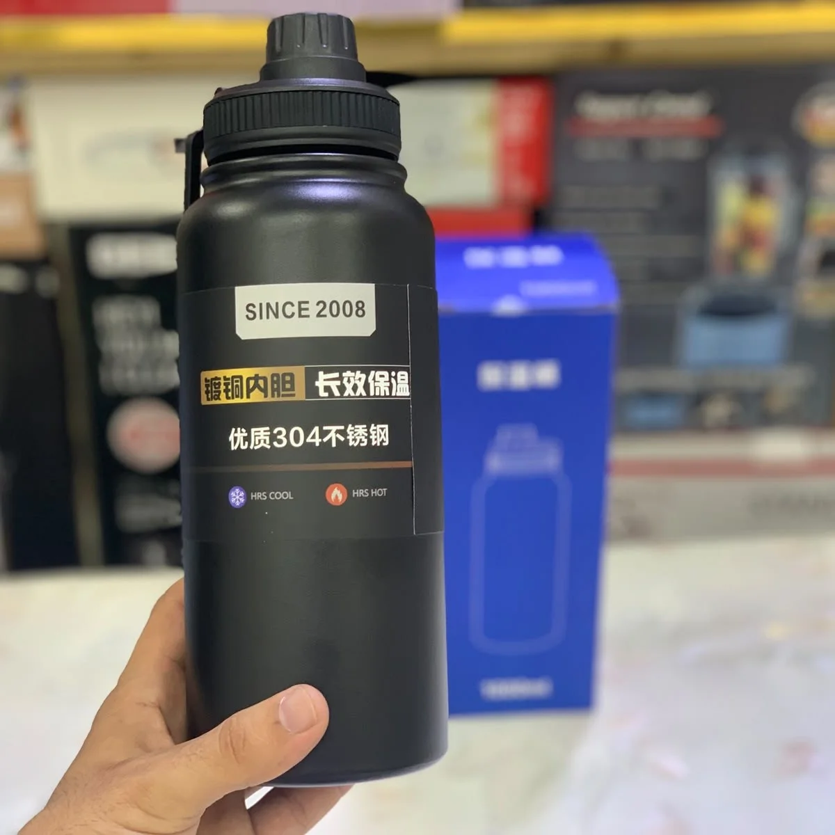 A black 1000ml insulated stainless steel vacuum water bottle with a carry handle and screw cap, placed beside its blue packaging box featuring Chinese text and a line illustration of the bottle.